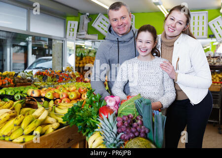 Cheerful family with preteen daughter standing with full grocery cart after shopping in fruit store Stock Photo
