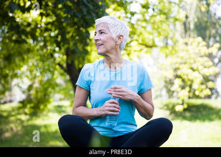 Senior woman rests and drinks water after workout in the park Stock Photo