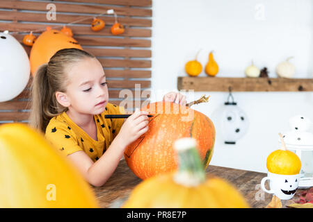 Cute young girl sitting at a table in living room, drawing a face on a large halloween pumpkin. Halloween holiday and family lifestyle background. Stock Photo