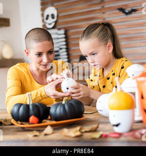 Cute young girl sitting at a table, decorating little white pumpkins with her mother, a cancer patient. DIY Halloween holiday and family lifestyle bac Stock Photo