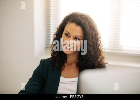 Close up of a smiling businesswoman in office. Curly haired businesswoman in formal clothes sitting at her desk in office. Stock Photo