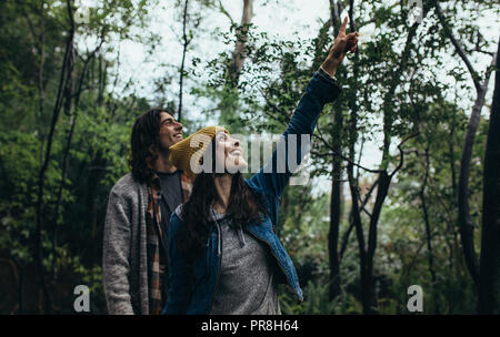 Young couple standing together in forest, with woman pointing up and smiling. Happy man and woman looking at something interesting in the forest. Stock Photo