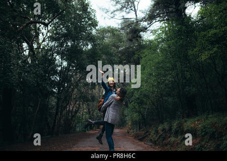 Man carrying her girlfriend raising hand and smiling. Couple enjoying themselves at forest on rainy day. Stock Photo