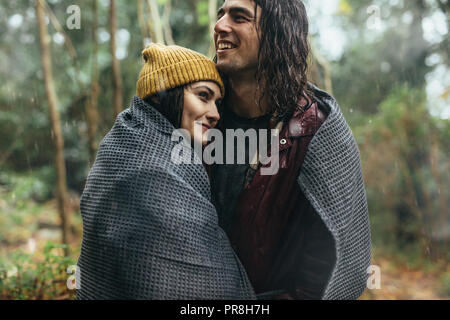 Beautiful young couple wrapped in a blanket on a rainy day. Caucasian couple embracing under the rain. Stock Photo