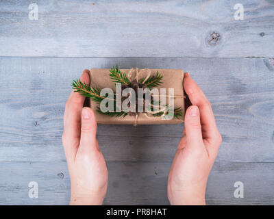 Female hands hold christmas holiday handmade present in craft paper with spruce branches over gray wooden table. Top view. Christmas background. Copy space Stock Photo