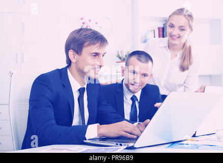 portrait of three smiling business colleagues occupied with laptops and chatting in office. Selective focus Stock Photo