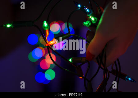 Female hand holding string of Christmas lights with defocused xmas tree in the background. Christmas lights bokeh lifestyle background. Stock Photo
