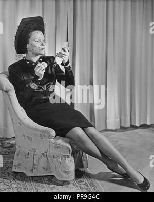 Swedish actress Margit Rosengren, 1901-1952. Pictured sitting in an armchair wearing a fashionable black dress, hat and veil. On her feet a pair of shiny black shoes and on her legs,  nylon stockings. She looks herself in the mirror while bettering the makeup.  Sweden 1945. Photo Kristoffersson Stock Photo