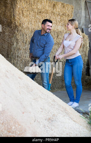 Smiling elderly man and young woman standing with metallic spades in hangar with sand pile Stock Photo
