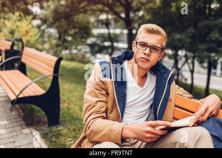 College student with backpack reading book walking in autumn park sitting on bench. Man studying outdoors