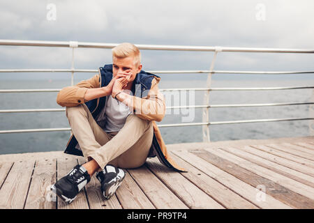 Upset young man sitting by sea on pier in Odessa thinking on rainy autumn day. Depression and stress concept. Mental health Stock Photo
