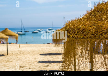 Straw parasols on a sandy beach with a yachts sailing on the background Stock Photo