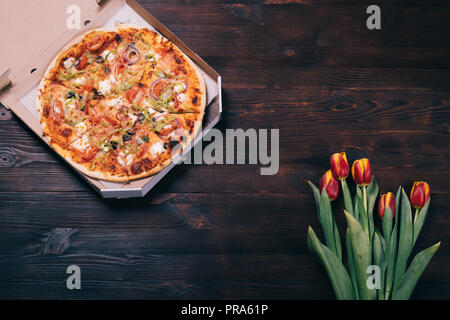 Round baked pizza in cardboard box on dark brown wooden table near bouquet of fresh red-yellow flowers tulips, top view. Stock Photo