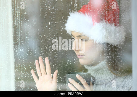 Sad and lonely woman looking through a window touching glass, with the hand in christmas time in a rainy day Stock Photo