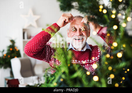 A senior man standing by Christmas tree, putting balls in front of eyes. Stock Photo