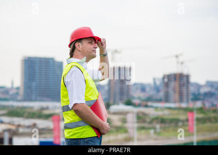 Engineer on a construction site wearing yellow reflective west and red helmet with buildings and cranes in the background. Foreman on a building site Stock Photo