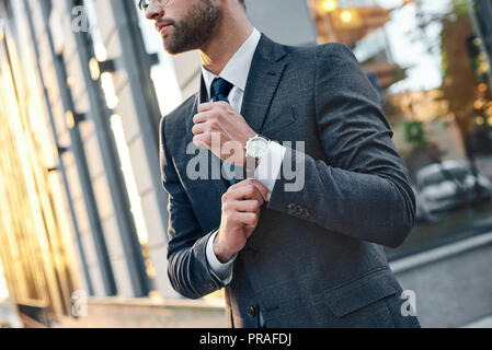 Cropped profile portrait of a successful young bearded guy in suit and glasses. So stylish and nerdy. Outdoors on a sunny street, fixing his cuffs Stock Photo