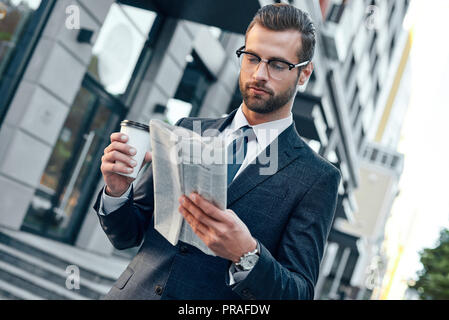 Young businessman in suit and glasses holding a paper cup and reading business newspaper in his hands Stock Photo