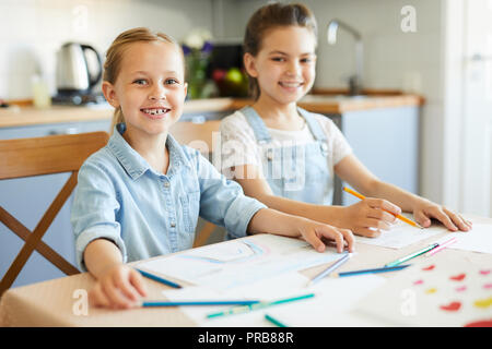 Two cheerful little girls in casualwear sitting by table in the kitchen while going to draw Stock Photo