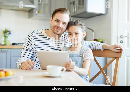 Affectionate young man and his little daughter sitting by table in the kitchen and watching online video Stock Photo