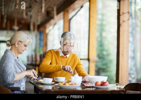 Content senior couple in casual clothing sitting at table and eating food while having breakfast at porch of countryside house Stock Photo