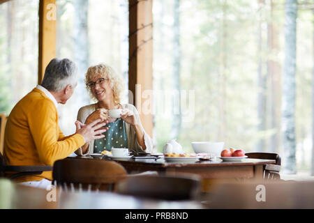 Positive beautiful mature couple in casual clothing sitting at table and drinking coffee at veranda while chatting in tranquil place Stock Photo