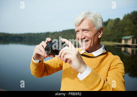 Smiling excited gray-haired senior tourist in yellow sweater taking photo of lake on camera and enjoying fresh air and beautiful nature Stock Photo