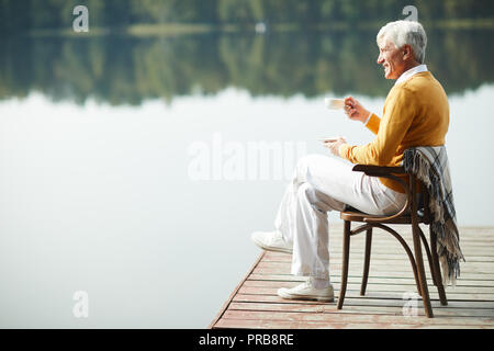 Happy relaxed handsome senior man wearing stylish clothing sitting on old chair with blanket on pier and drinking coffee while contemplating lake Stock Photo