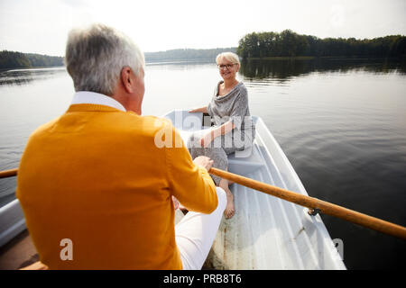 Happy attractive elderly lady in dress and glasses sitting on boat and looking at man while they riding boat during beautiful date on lake Stock Photo
