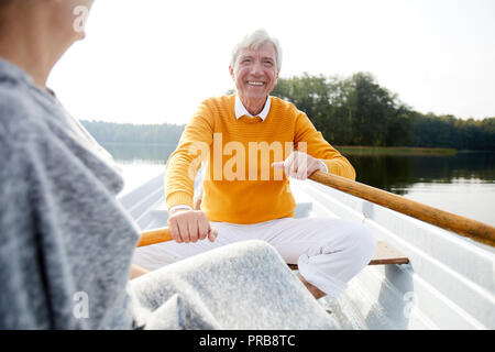Cheerful excited handsome elderly man in shirt and yellow sweater sitting on boat and rowing with oars while talking to woman Stock Photo