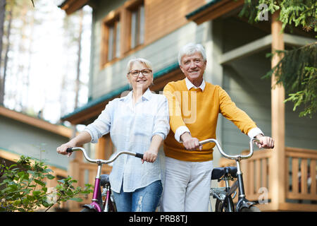 Cheerful active senior couples in casual clothing standing against country house and holding bicycles while looking at camera Stock Photo
