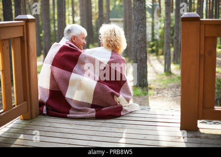 Rear view of happy senior couple covered with checkered blanket talking and sitting on porch of country house Stock Photo