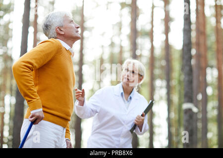Rehabilitation specialist in glasses supporting senior patient and talking to him while they walking in forest, motivated senior man learning to walk  Stock Photo