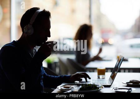 Silhouette of male hipster entrepreneur in headphones sitting at table and using laptop while drinking coffee and working in cafe Stock Photo