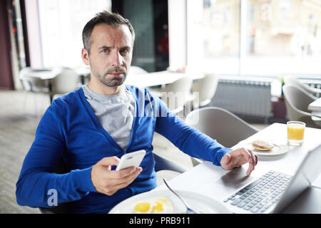 Serious concentrated businessman in blue cardigan sitting at table and using smarphone while answering sms, he telecommuting in cafe Stock Photo