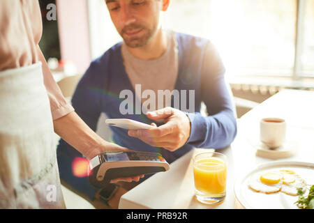 Close-up of handsome man paying bill through smartphone and putting device to cash terminal held by waitress in cafe Stock Photo