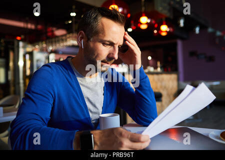 Serious thoughtful handsome businessman in earbuds concentrated on text information reading contract while sitting in cafe Stock Photo