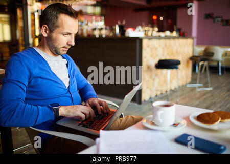 Content enterprising handsome male freelancer in blue cardigan sitting at table and keeping laptop on knees while doing work in cafe during breakfast Stock Photo