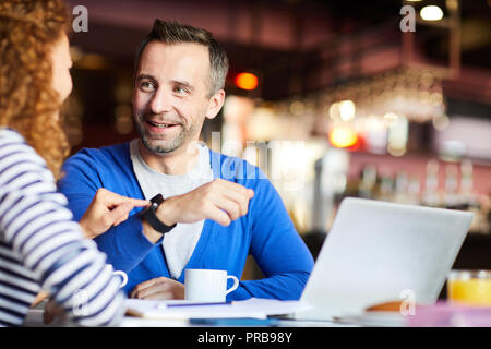 Cheerful confident handsome man with stubble showing new smartwatch to colleague who touching screen and viewing function of device in cafe Stock Photo