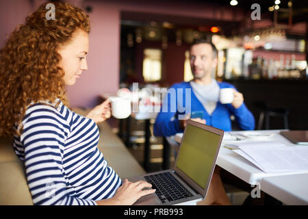 Coworking space for business: serious pensive female entrepreneur with curly hair drinking coffee and browsing internet on laptop Stock Photo