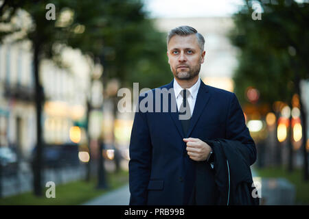 Mature serious businessman in elegant suit holding his coat while walking in urban environment Stock Photo