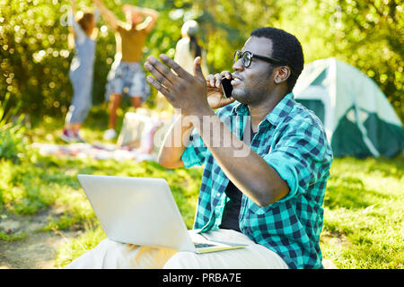 African-american guy in casualwear talking by smartphone on backpack trip with his friends on background Stock Photo