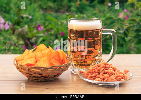Glass mug of beer on wooden table with potato chips in wicker basket, peanuts in plate on natural green background Stock Photo