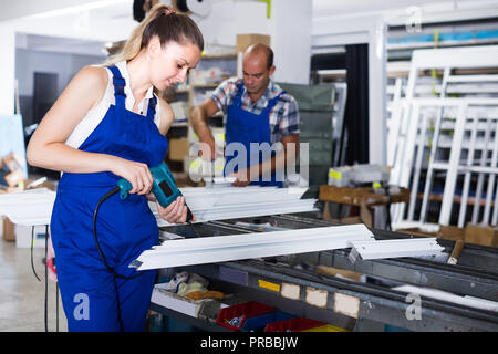 Female worker is assembling metal-plastic window in workshop, using electric drill. Stock Photo