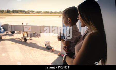 Toned image of young mother holding her toddler son and showing airplanes on runway through big window in airport terminal Stock Photo