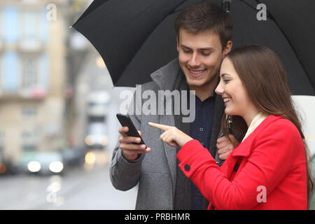 Happy couple checking smart phone content holding an umbrella in winter under the rain Stock Photo