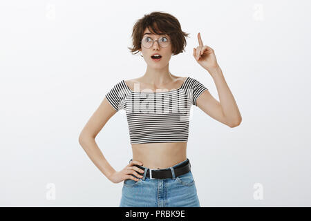 I know what to do. Portrait of good-looking excited woman in round glasses and striped cropped top, raising index finger in eureka gesture and standing with opened mouth, having suggestion Stock Photo