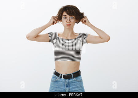 Portrait of playful childish cute woman in round eyewear and cropped striped top, pulling ears and making funny face, pouting, standing carefree and bored over gray background. having fun Stock Photo