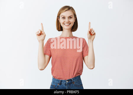 Creative and happy girl going only forward. Charming caucasian girlfriend with fair hair in striped t-shirt, pointing up with index fingers and smiling broadly with satisfied optimistic expression Stock Photo