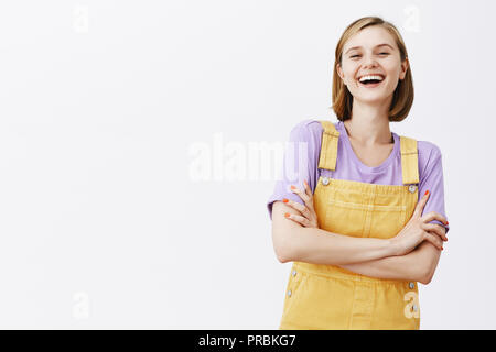 Carefree caucasian girl with fair hair in yellow overalls, holding hands crossed on chest in self-assured pose, laughing out loud and smiling at camera, having fun, talking with coworkers during break Stock Photo